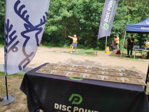 Na starcie dołka do disc golfa stoi osoba w pomarańczowej koszulce. Widok zza stolika na którym leżą rzędy drewnianych nagród.