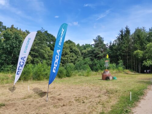Kosz do disc golfa wyniesiony na drewnianym pieńku. Po jego lewej stronie biało-niebieskie flagi z napisem „PDGA Europe”. Po prawej stronie w oddali widać grupę grających osób.