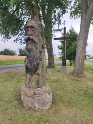 Przypominająca pogański posąg spora drewniana rzeźba brodacza z wybałuszonymi oczami i otwartymi ustawi stoi przy przydrożnej kępie drzew, pomiędzy którymi stoi również drewniany krzyż.