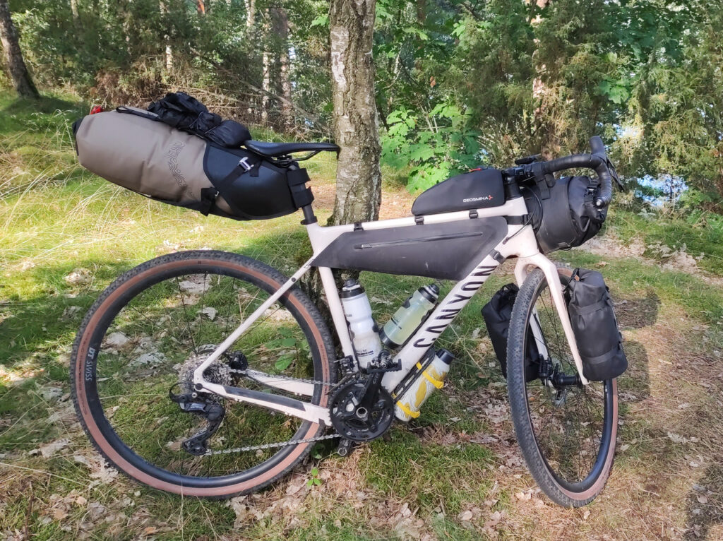 Mój jasnokremowy, prawie biały rower szutrowy Canyon Grizl, zwany dalej Gryzelkiem, stoi oparty o drzewko. Z tyłu pod siodłem ma duży jasnobrązowy worek wpięty w czarną uprząż. Wewnątrz ramy, pod górną rurą znajduje się ciasno dopasowana czarna torba, a pod nią dwa bidony. Na ramie przy kierownicy jest czarna torba na podręczne żarcie. Pod ramą przypięta żółtymi paskami elastycznymi do czarnego koszyka jest metalowa butelka na wodę. Na widelcu w czarnych koszykach są wypchane worki. Na kierownicy kolejna uprząż ze zwiniętym z obu stron czarnym workiem.