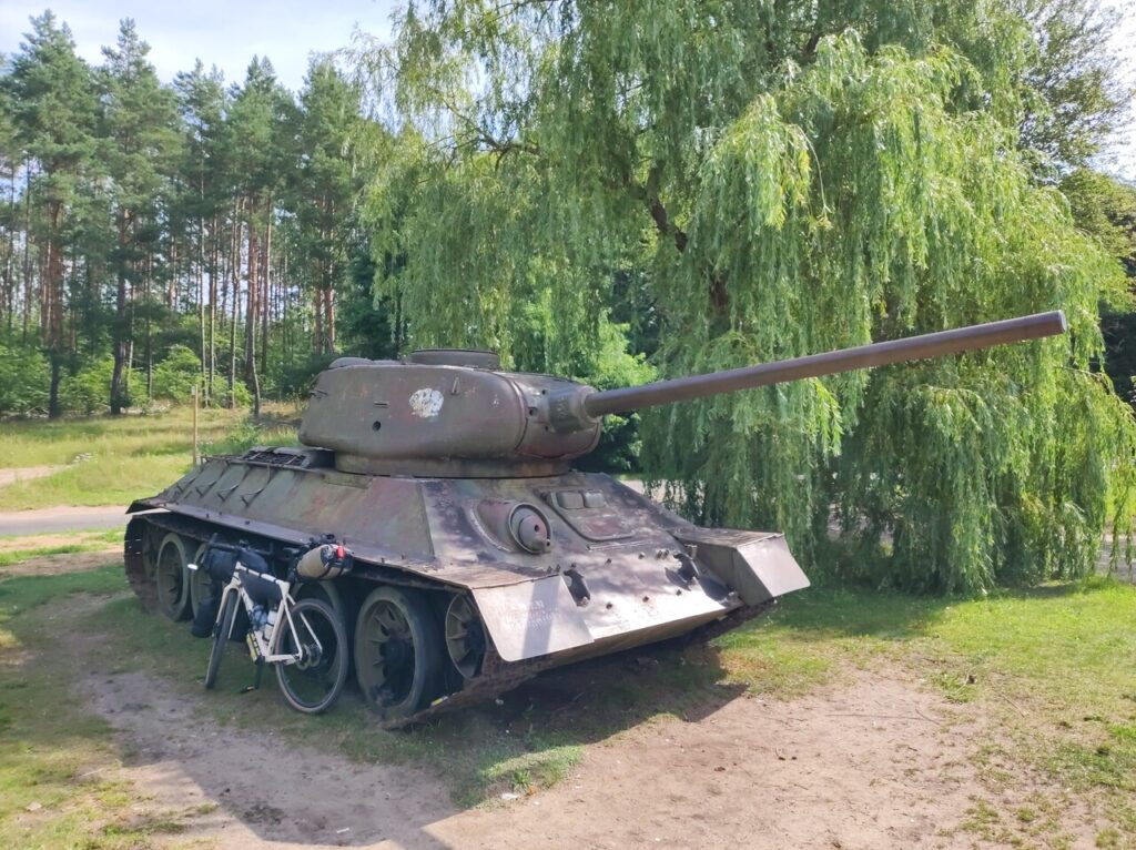 Zwrócony w prawo czołg T-34/85 i oparty o prawą gąsienicę rower szutrowy z bikepackingowymi torbami. Za nim po prawej jest wierzba ze zwieszającymi się gałęziami, w dali po lewej rzadki sosnowy las.