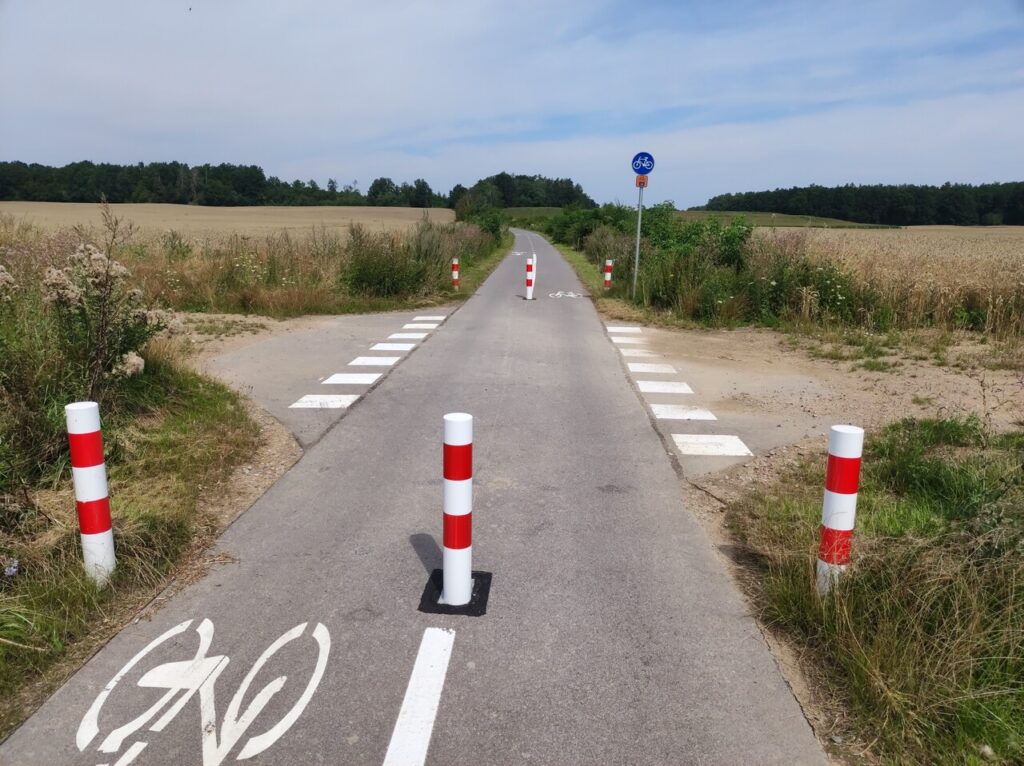 Skrzyżowanie z polną drogą na Starym Kolejowym Szlaku. Asfaltowa droga dla rowerów jest z obu stron przegrodzona trzema biało-czerwonymi słupkami. Za skrzyżowaniem po prawej stoi niebieski znak C-13 z białym symbolem roweru, pod którym jest pomarańczowy kwadrat z oznaczeniem trasy numer 15.
