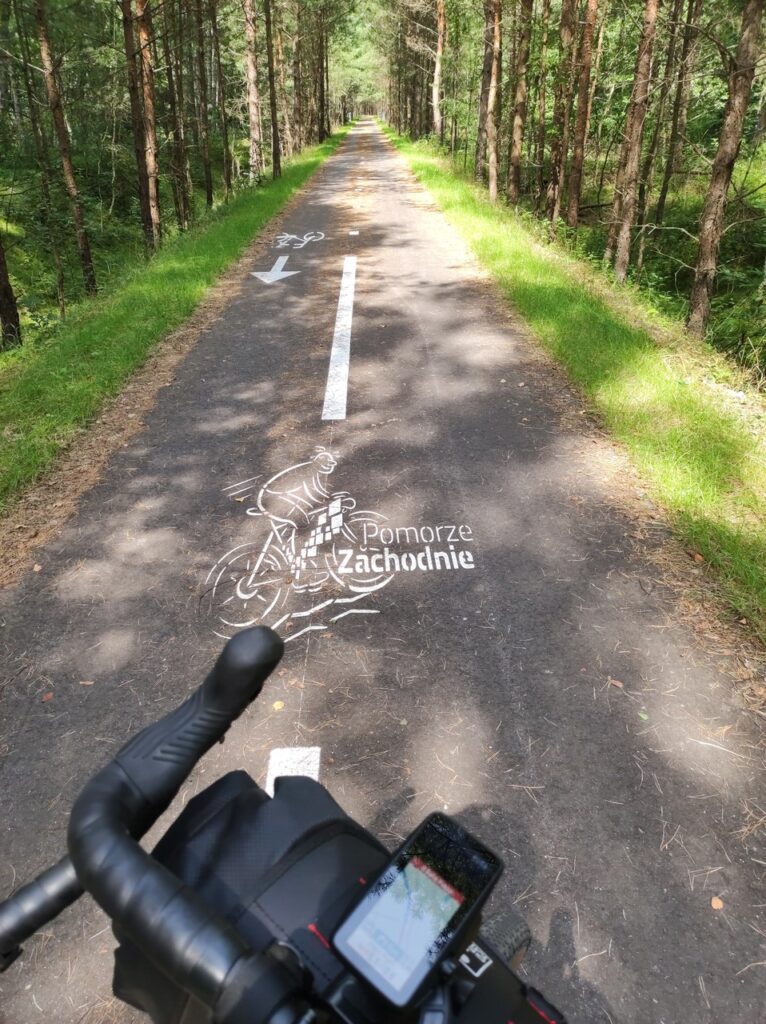 Asfaltowa droga dla rowerów przedzielona na pół białym pasem. Pomiędzy pasami rysunek osoby na rowerze z dopiskiem „Pomorze Zachodnie”.