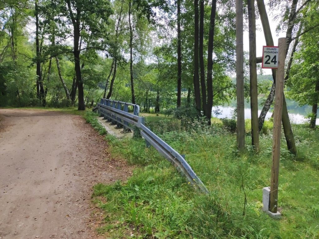 Droga szutrowa biegnąca przez mostek nad niedużym ciekiem. Po prawej metalowa barierka, słupek oznaczający dojazd pożarowy 24, a między drzewami widać wodę jeziora.