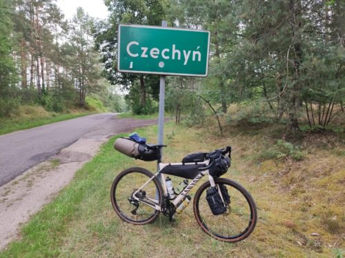 Pobocze wąskiej asfaltowej drogi. O znak z nazwą miejscowości Czechyń opiera się w pełni objuczony Gryzelek. Dookoła drogi las.