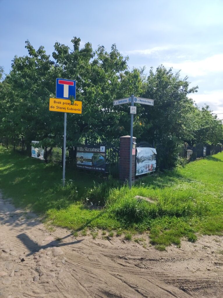 Róg ulic Garncarza i Wojciecha Brezy w Skrzatuszu. W prawo, na ulicę Brezy pokazuje oznaczenie zielonego szlaku rowerowego. Na początku ulicy Garncarza stoi znak „droga bez przejazdu” z żółtą tabliczką „Brak przejazdu do Starej Łubianki”
