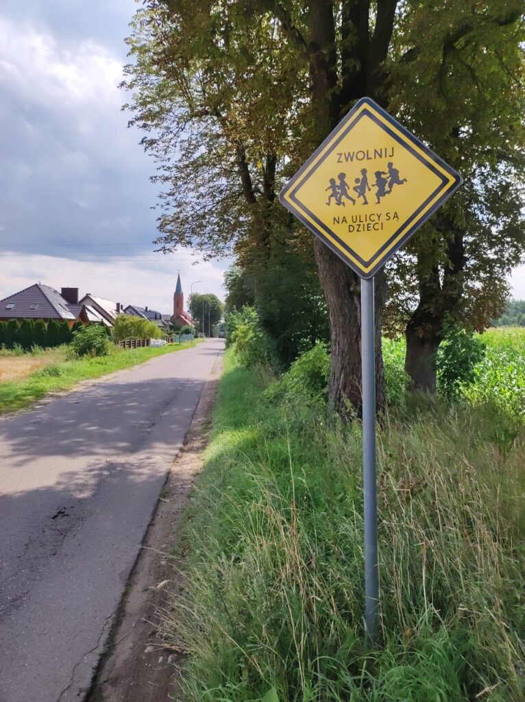Przy wąskiej szosie z podniszczonym asfaltem stoi znak w postaci żółtego trójkąta ustawionego na jednym z rogów z napisem „Zwolnij, na ulicy są dzieci” i rysunkiem sylwetek bawiących się dzieci.