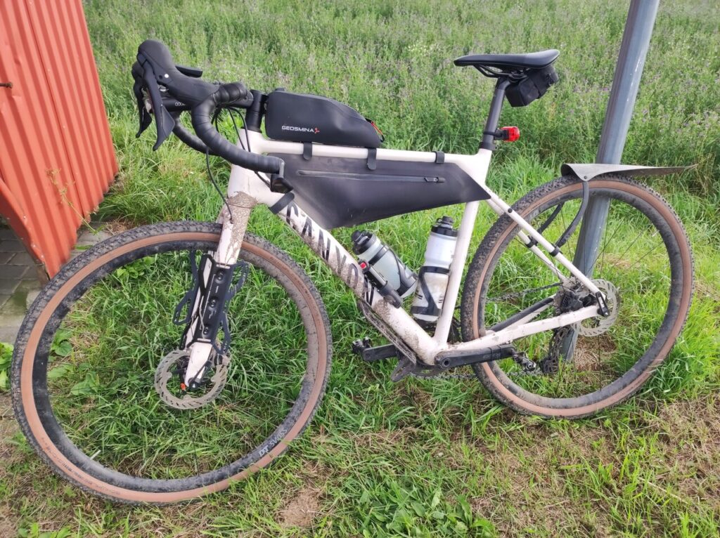 Mój jasnokremowy, prawie biały rower szutrowy Canyon Grizl oparty tylnym kołem o słupek znaku drogowego. Rower jest brudny od zaschniętego błota. W ramie jest czarna torba oraz dwa białe bidony.