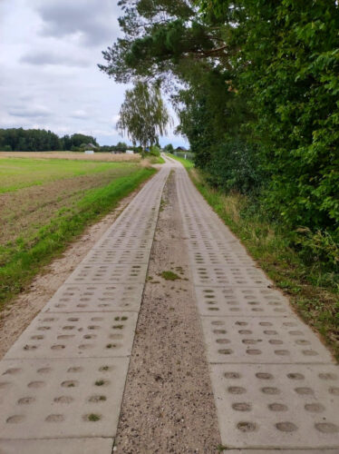 Prosta droga z polem po lewej stronie i drzewami po prawej, wyłożona ażurowymi płytami.