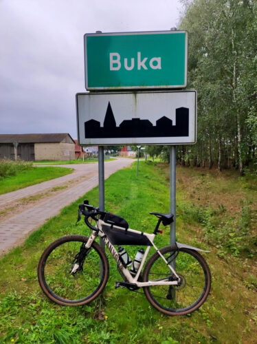O znak z nazwą miejscowości Buka opiera się mój rower. Za nim po prawej lasek brzozowy, a po lewej zabudowania gospodarstwa.