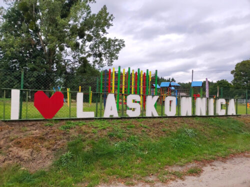 Wycięty ze styropianowych płyt napis I [serce] LASKOWNICA stojący przy płocie, za którym znajduje się przyszkolny plac zabaw.